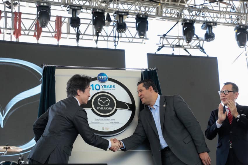 Guanajuato governor Diego Sinhue shaking hands with a Japanese Mazda official at a ceremony. Behind them is a sign made to look like a car driving wheel that says GTO, 10 years and Mazda in large type.