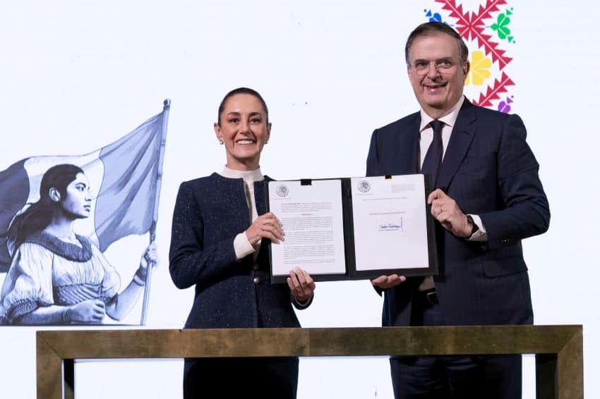 Mexico's President Claudia Sheinbaum and its Economy Minister Marcelo Ebrard stand at a press conference in front of a long, thin table. Together they are holding up a portfolio cover with the document they signed to expand a 35% tariff on textile imports.