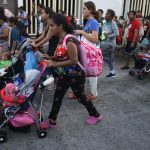 A dark-skinned young woman pushing a child in a stroller leads a group of migrants down a Mexican street, part of a migrant caravan