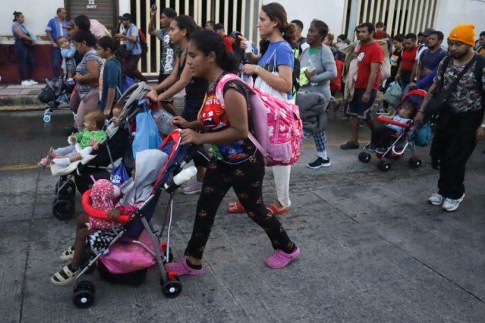 A dark-skinned young woman pushing a child in a stroller leads a group of migrants down a Mexican street, part of a migrant caravan