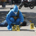 A forensic expert in blue protective gear and wearing a surgical mask and gloves in a city street in Chilpancingo puts down two yellow plastic marker signs on the ground to mark evidence in a crime scene. A dead victim lies sprawled on the sidewalk behind the expert.