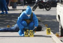 A forensic expert in blue protective gear and wearing a surgical mask and gloves in a city street in Chilpancingo puts down two yellow plastic marker signs on the ground to mark evidence in a crime scene. A dead victim lies sprawled on the sidewalk behind the expert.