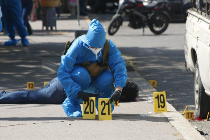 A forensic expert in blue protective gear and wearing a surgical mask and gloves in a city street in Chilpancingo puts down two yellow plastic marker signs on the ground to mark evidence in a crime scene. A dead victim lies sprawled on the sidewalk behind the expert.