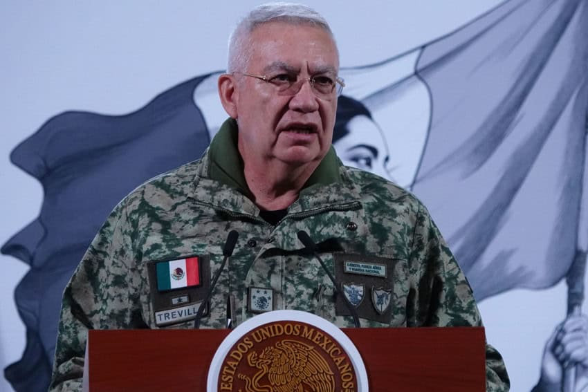 Mexico's Defense Minister Ricardo Trevilla standing at a podium at President Sheinbaum's daily press conference, speaking to reporters. He has a gray-haired receding hairline, is dressed in military fatigues, and wears wire-rimmed glasses.