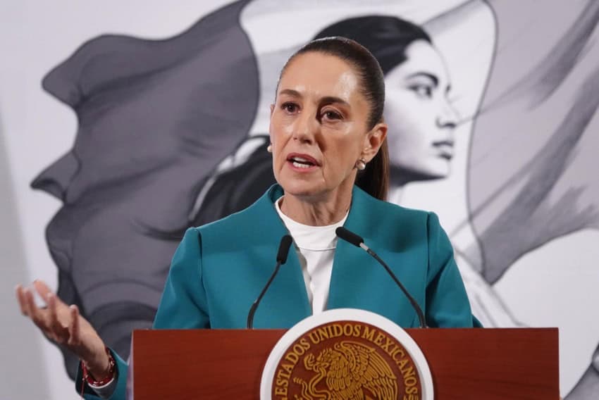 Mexican President Claudia Sheinbaum gesturing with her right hand as she speaks at the presidential podium during a press conference. She's wearing a turqouise blazer with a white shirt underneath. Behind her is the symbol of her administration, a young Indigenous woman holding the Mexican flag