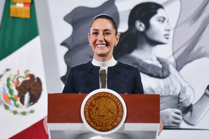 Mexico's President Claudia Sheinbaum at the presidential podium in the National Palace. She is smiling while responding to reporters during her press conference.