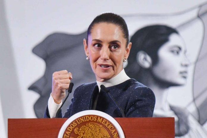 Claudia Sheinbaum in mid speech while standing at the presidential podium during a press conference. She is looking forward with her gaze pointed toward her left and is holding her right hand up before her chest in a closed fist.