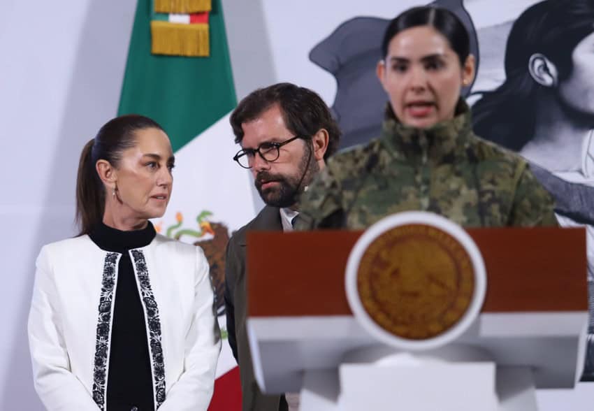 A woman in military fatigues speaks at Mexico's presidential podium during a presidential press conference while President Claudia Sheinbaum confers in the background onstage with Mexican Social Security director Alejandro Svarch.