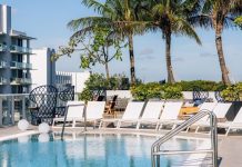 Image of a luxury hotel in a tropical location, with a rooftop pool with white deck chairs around it, palm trees behind it and buildings in the far left background with wide balconies on each floor, like a hotel.