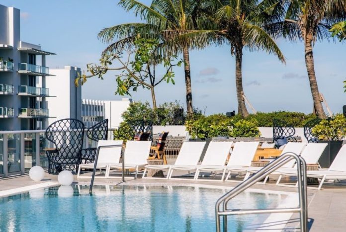 Image of a luxury hotel in a tropical location, with a rooftop pool with white deck chairs around it, palm trees behind it and buildings in the far left background with wide balconies on each floor, like a hotel.