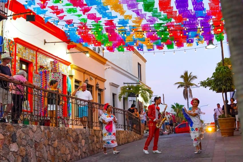 Tourists watch musicians perform in the street under colorful paper flags next to a colonial building