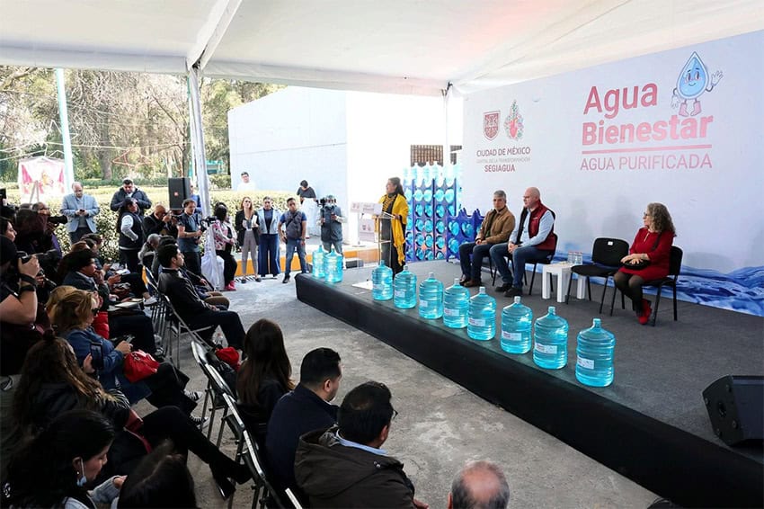 Mexico City Mayor Clara Brugada and other officials on stage with 20-liter jugs of water and a banner reading "Agua Bienestar"