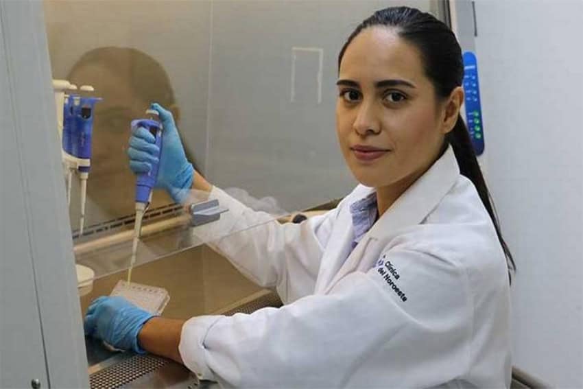 Young woman in a lab coat and scientific lab research equipment in her hands. She is wearing latex gloves and staring at the camera