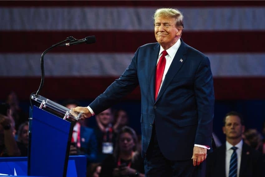 Donald Trump standing sideways next to a podium on a stage in a stadium. In the background is a crowd of spectators and behind them a giant US flag that's not visible in full in the photo due to its size