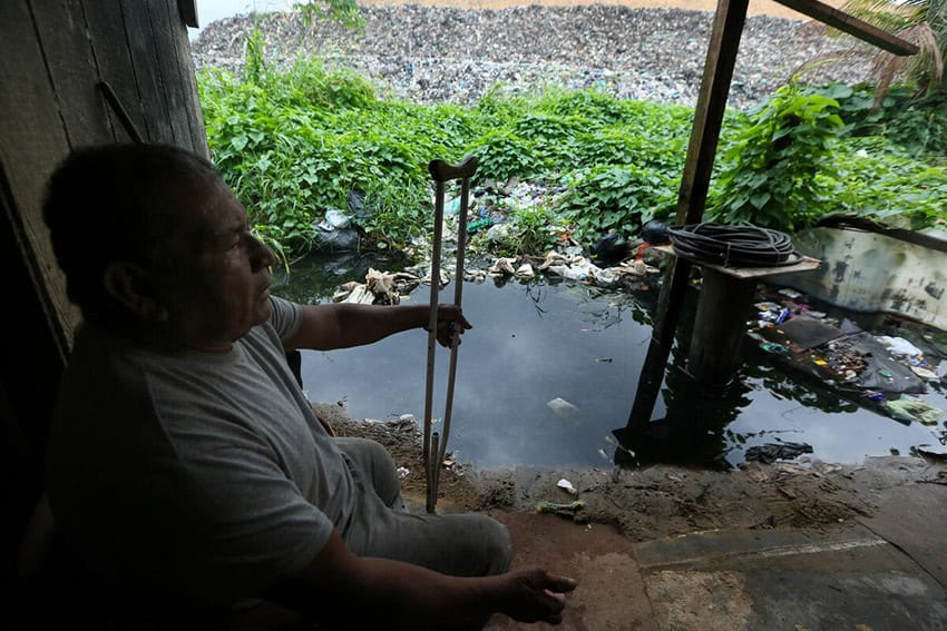 A man with a crutch sits next to a small pond filled with plastic waste, in front of a trash dump.