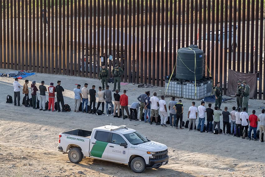 A group of migrants, mostly men, line up in front of two border agents in green uniforms near the border wall on June 6, two days after Biden issued the executive order.