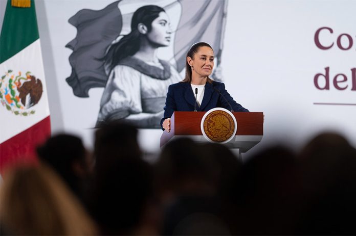 President Claudia Sheinbaum stands at a podium next to a Mexican flag at her morning press briefing