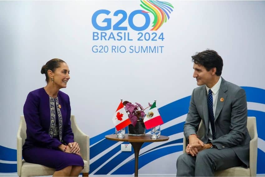 Claudia Sheinbaum and Justin Trudeau smiling and talking as they sit in white upholstered chairs side by side. facing opposite each other. They have a small wooden table between them with a tiny Mexican flag and Canadian flag. Each leader has the other leader's flag on their side. Behind them is a wall with the logo for the 2024 G20 Leaders' Summit