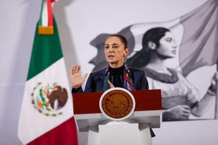 Claudia Sheinbaum standing at the presidential podium at the National Palace at her daily press conference. Her left hand is raised with her palm facing reporters as she speaks.