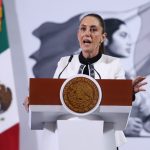 Mexico's President Claudia Sheinbaum standing at the presidential podium during her press conference. Her mouth is open in speech and her eyes are wide and round and she's gripping the podium.