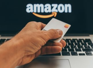 A hand holding a blank Master Card credit card in front of a laptop screen with the Amazon logo in white and orange.