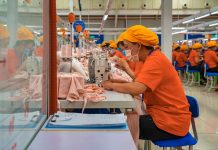 Female employees of a textile factory in Mexico. The women are at work, sitting in rows at tables with industrial sewing machines. They are wearing orange t-shirts and matching orange hats under which their hair is tucked.