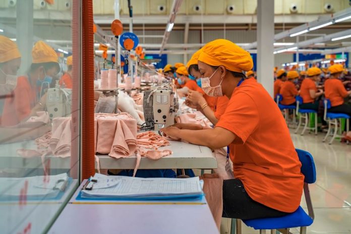 Female employees of a textile factory in Mexico. The women are at work, sitting in rows at tables with industrial sewing machines. They are wearing orange t-shirts and matching orange hats under which their hair is tucked.