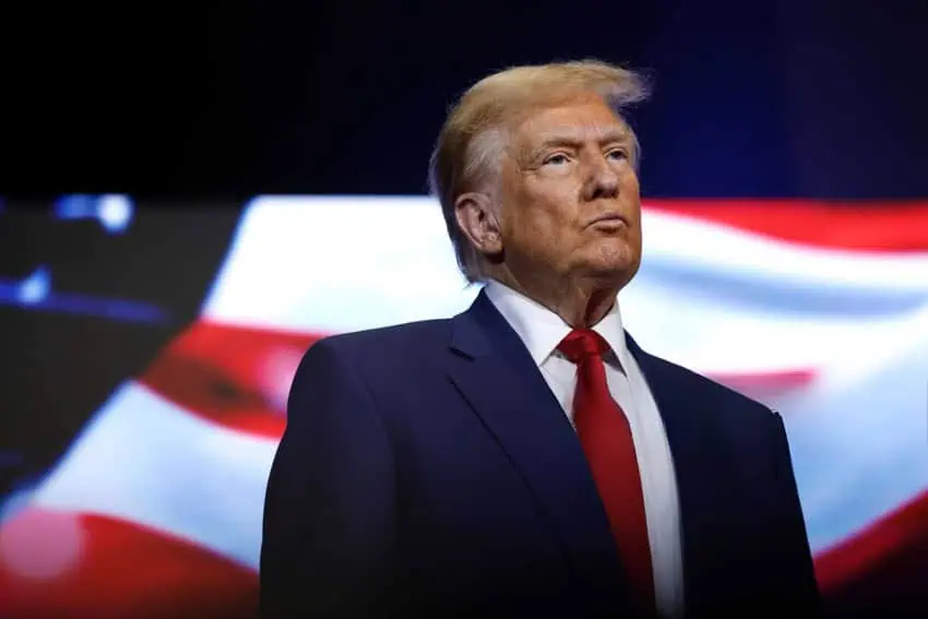 Photo of Donald Trump in a suit and tie standing in front of a projection screen of a rippling U.S. flag. His lips are pursed and he appears to be listening to someone speaking.