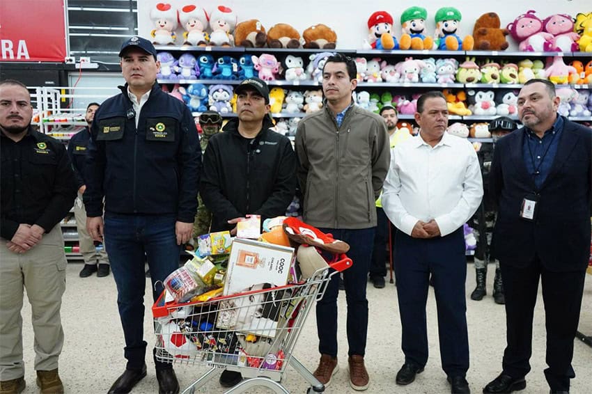 Sonora state officials stand in a row behind a cart full of small, colorful products, in front of a shelf full of stuffed toys