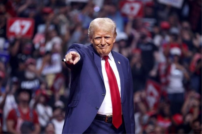 Donald Trump in a blue suit and red tie at a 2024 campaign rally, standing onstage in a stadium filled with people, smiling closemouthed as he points out into the audience