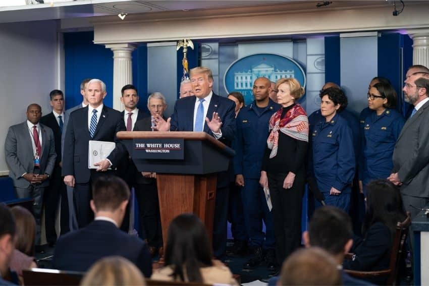 Donald Trump at the White House podium during a press conference, gesturing with his hands spread in front of him. He is surrounded by Vice President Pence and many other high-ranking government officials onstage